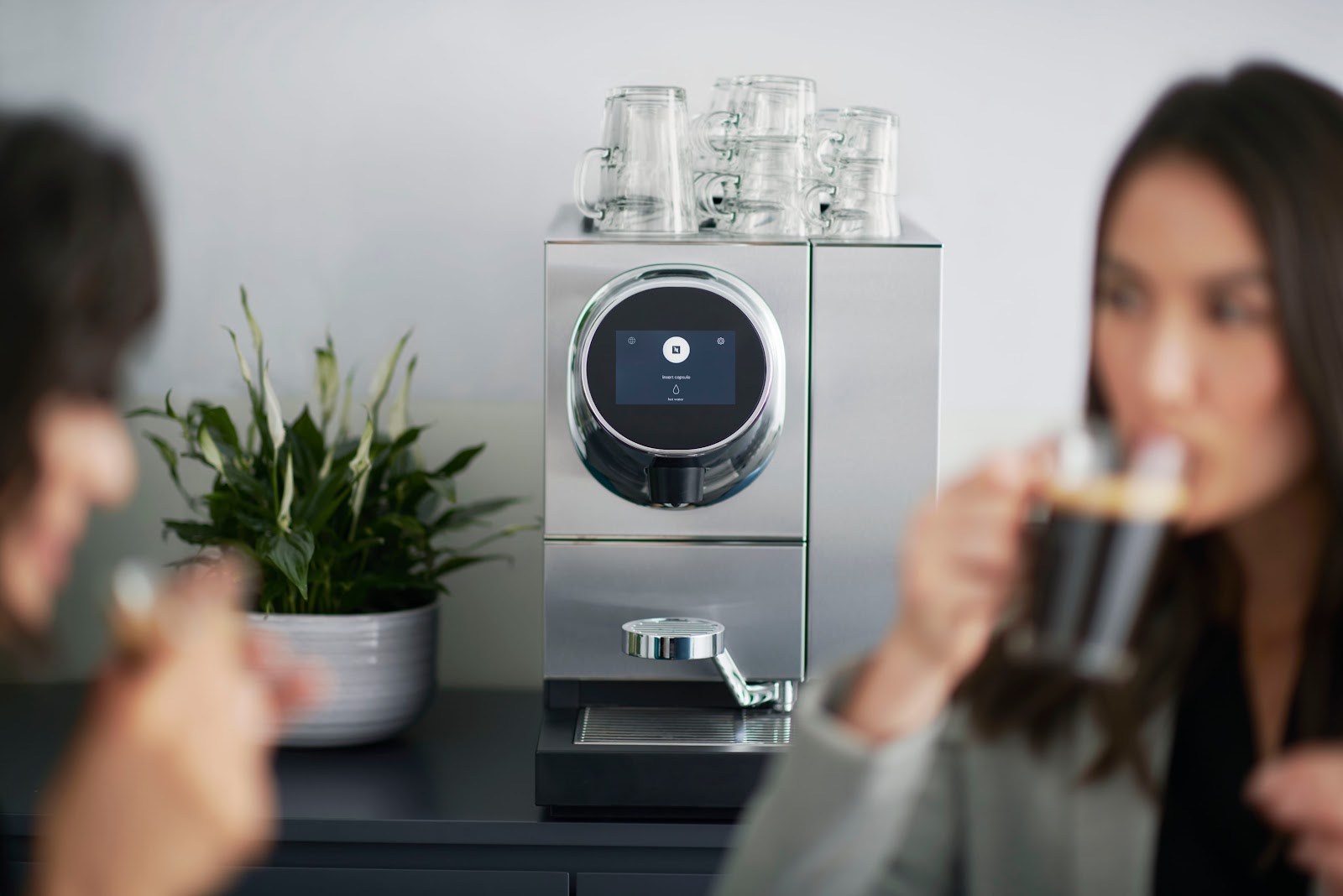 Two women drinking coffee next to a silver Nespresso coffee machine and a plant.