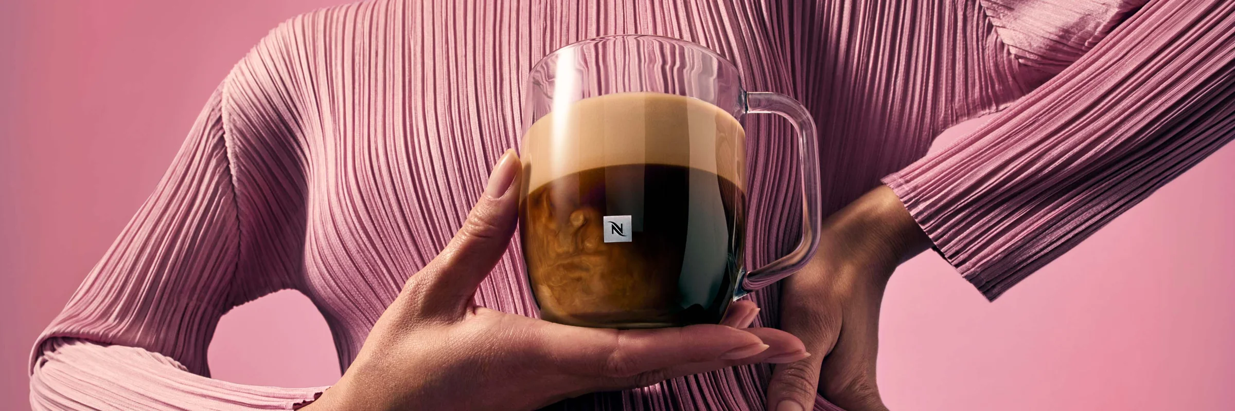 Person holding a glass Nespresso coffee mug against a pink background